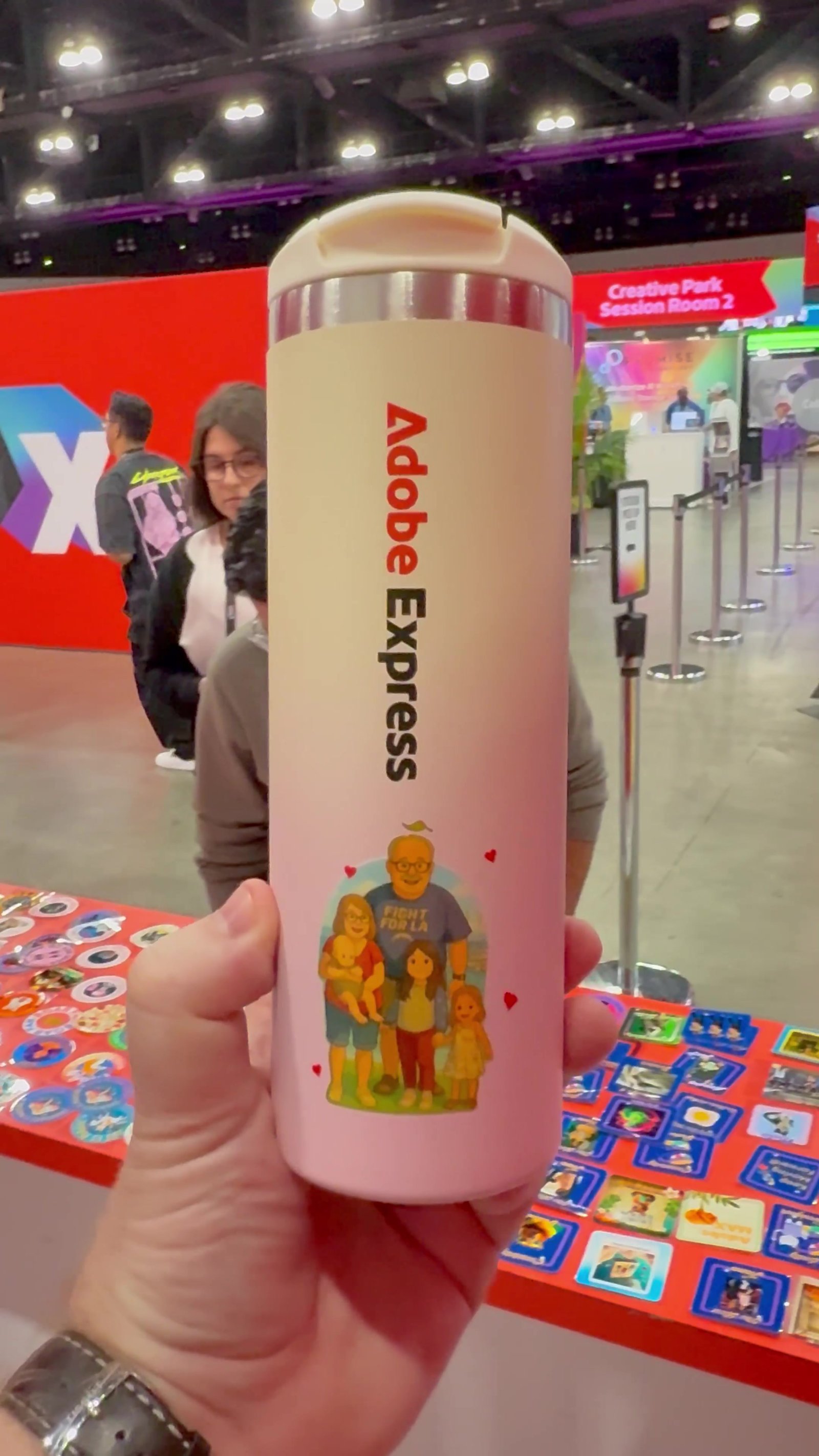 Full-color printed tumbler held up at a convention with a sticker station in the background